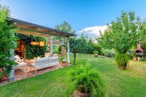 A covered patio with outdoor seating, a brick wall, and lush greenery. - Villa Orentano in Orentano (Castelfranco di Sotto)