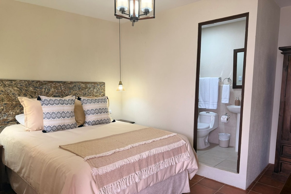 Un dormitorio con cama, almohadas, lámpara de noche, espejo y vista al baño.