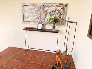 Una habitación con piso de baldosas, una pequeña mesa con un florero y un cuadro enmarcado, además de una figura decorativa de un caballo.