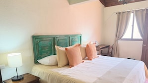 Uma cama com edredom branco e duas almofadas estampadas, uma mesa de cabeceira de madeira com abajur e uma pequena mesa com uma garrafa.