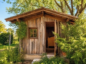 Una cabana de fusta rústica amb una única porta i finestra, envoltada de vegetació.