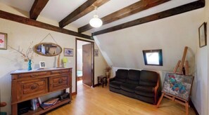 Een loft-achtige kamer met zichtbare houten balken, een zwarte bank, een houten dressoir en een schildersezel met een schilderij.