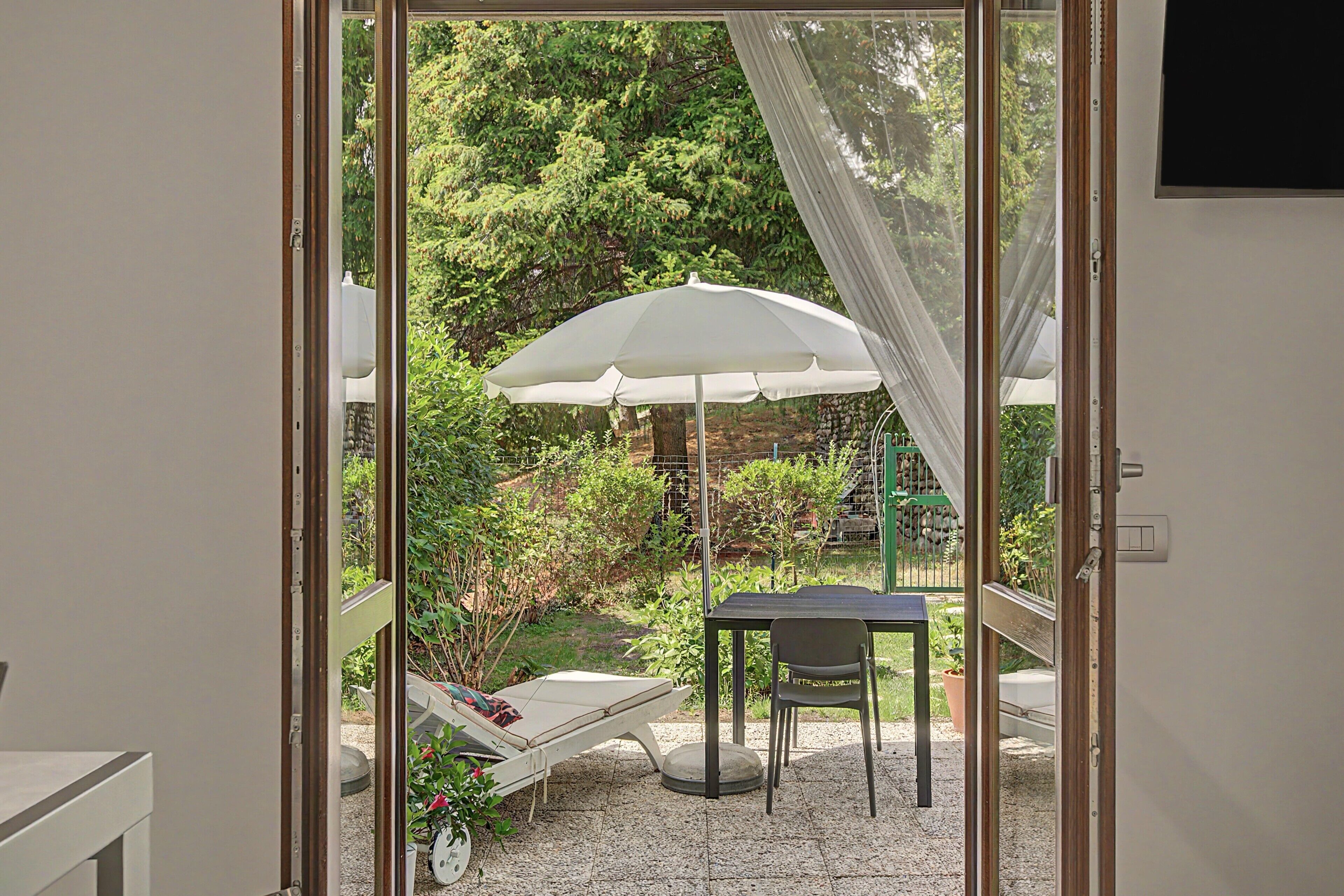 A room with a glass door leading to an outdoor area featuring a white umbrella, a lounge chair, and a table.