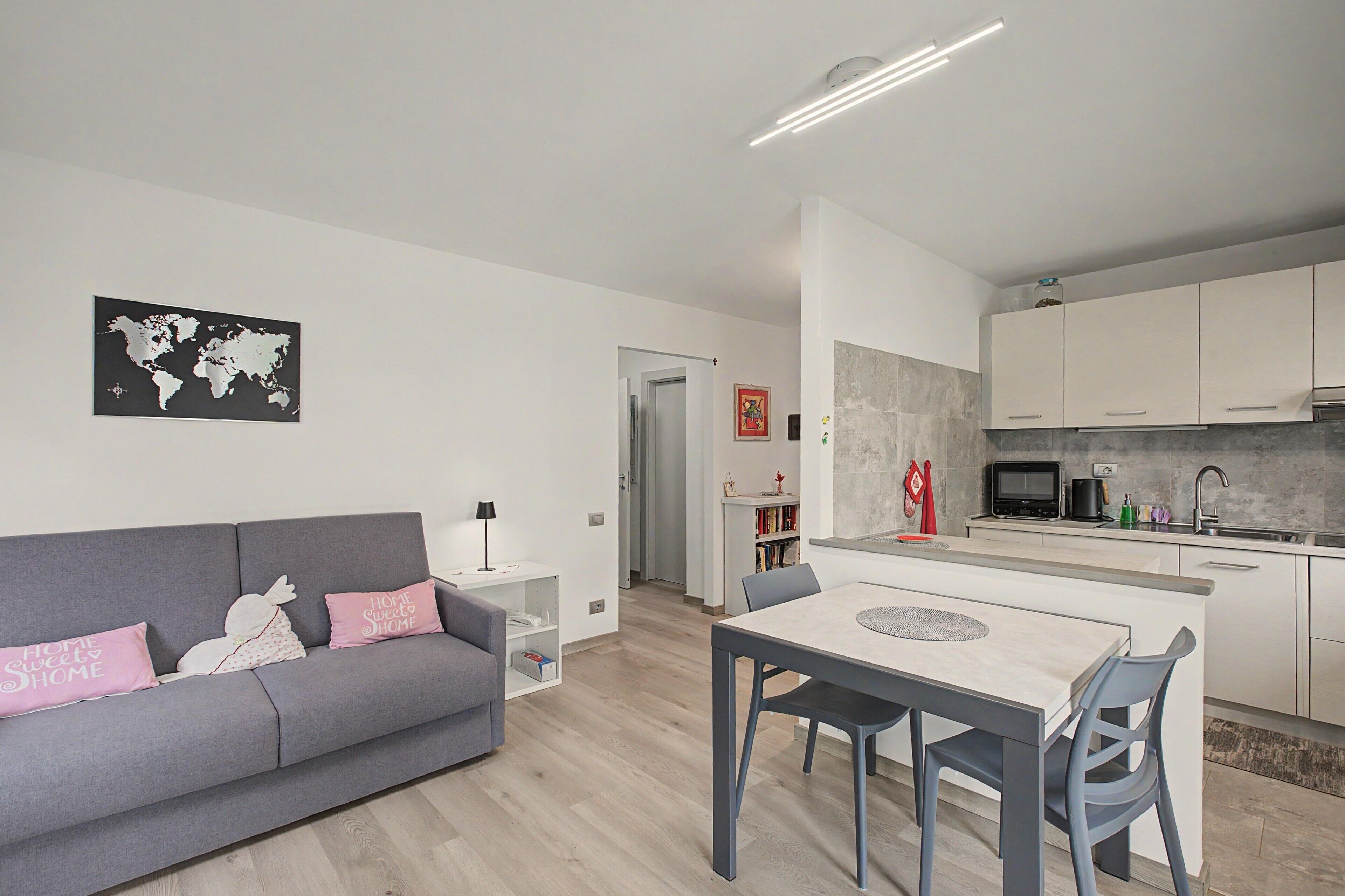 Un apartamento moderno con un sofá gris, una mesa de comedor con sillas, una cocina con armarios blancos y un mapa mundial en la pared.