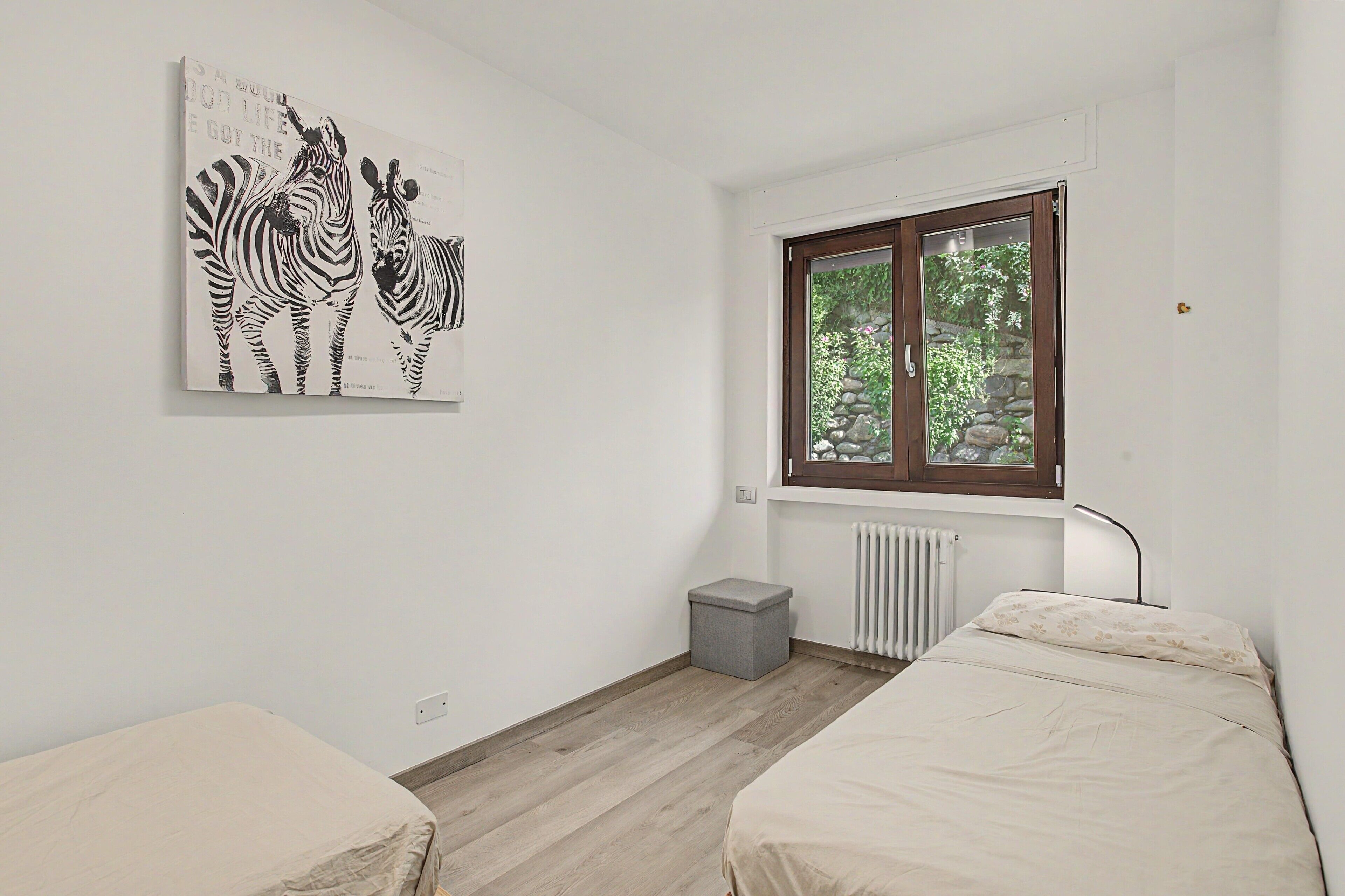 Quarto com duas camas, janela com vista para área verde, radiador e uma obra de arte com tema de zebra na parede.