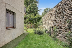 Une petite cour bordée d’un mur de pierre, avec une zone herbeuse et un bâtiment doté d’une fenêtre.