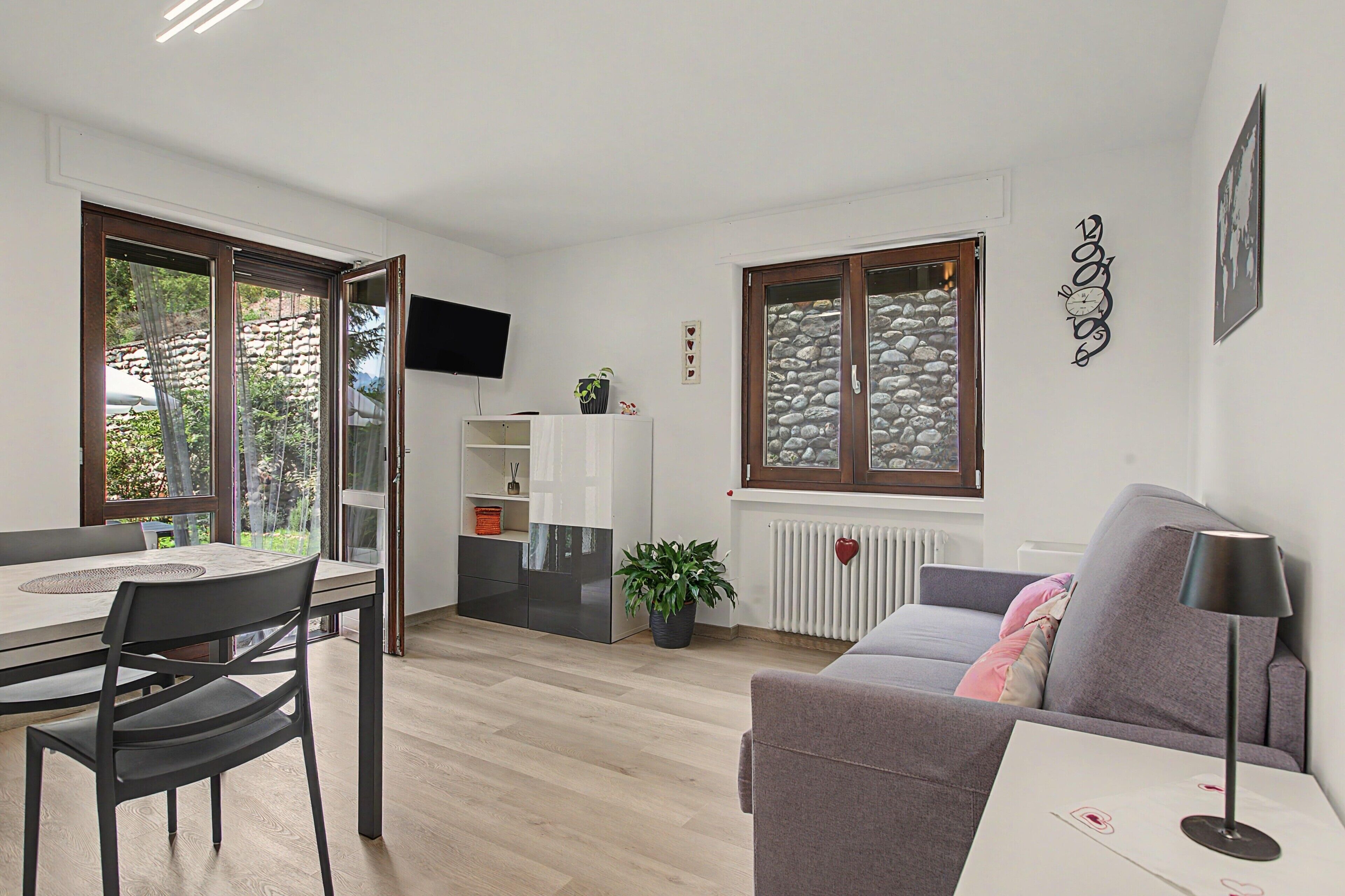 Una sala de estar moderna con comedor, un sofá gris, una mesa con sillas, un televisor montado en la pared y una ventana con fachada de piedra.
