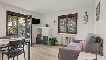 Una sala de estar moderna con comedor, un sofá gris, una mesa con sillas, un televisor montado en la pared y una ventana con fachada de piedra.