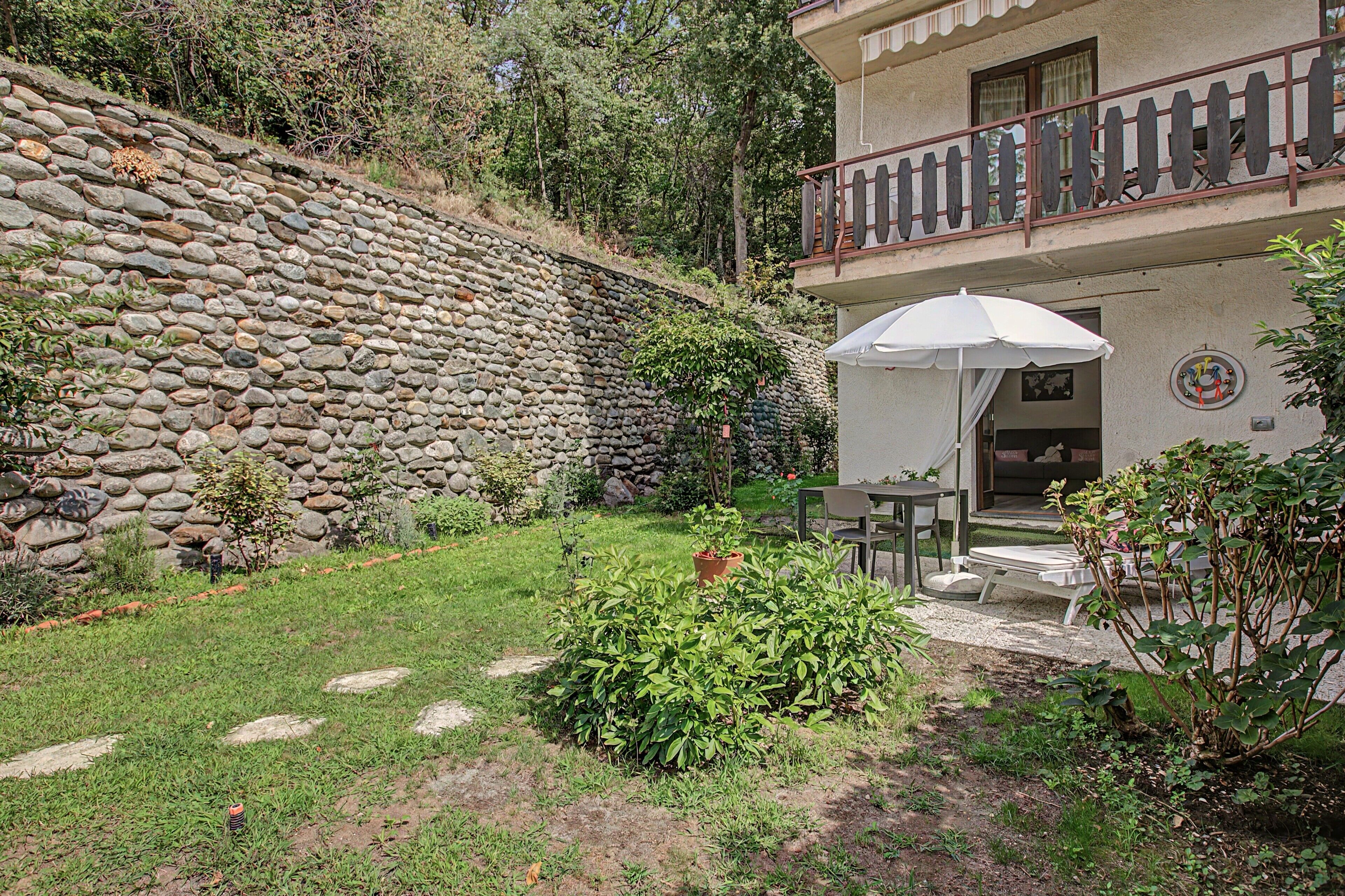Um muro de pedra, um jardim com mesa e cadeiras e um edifício com varanda.