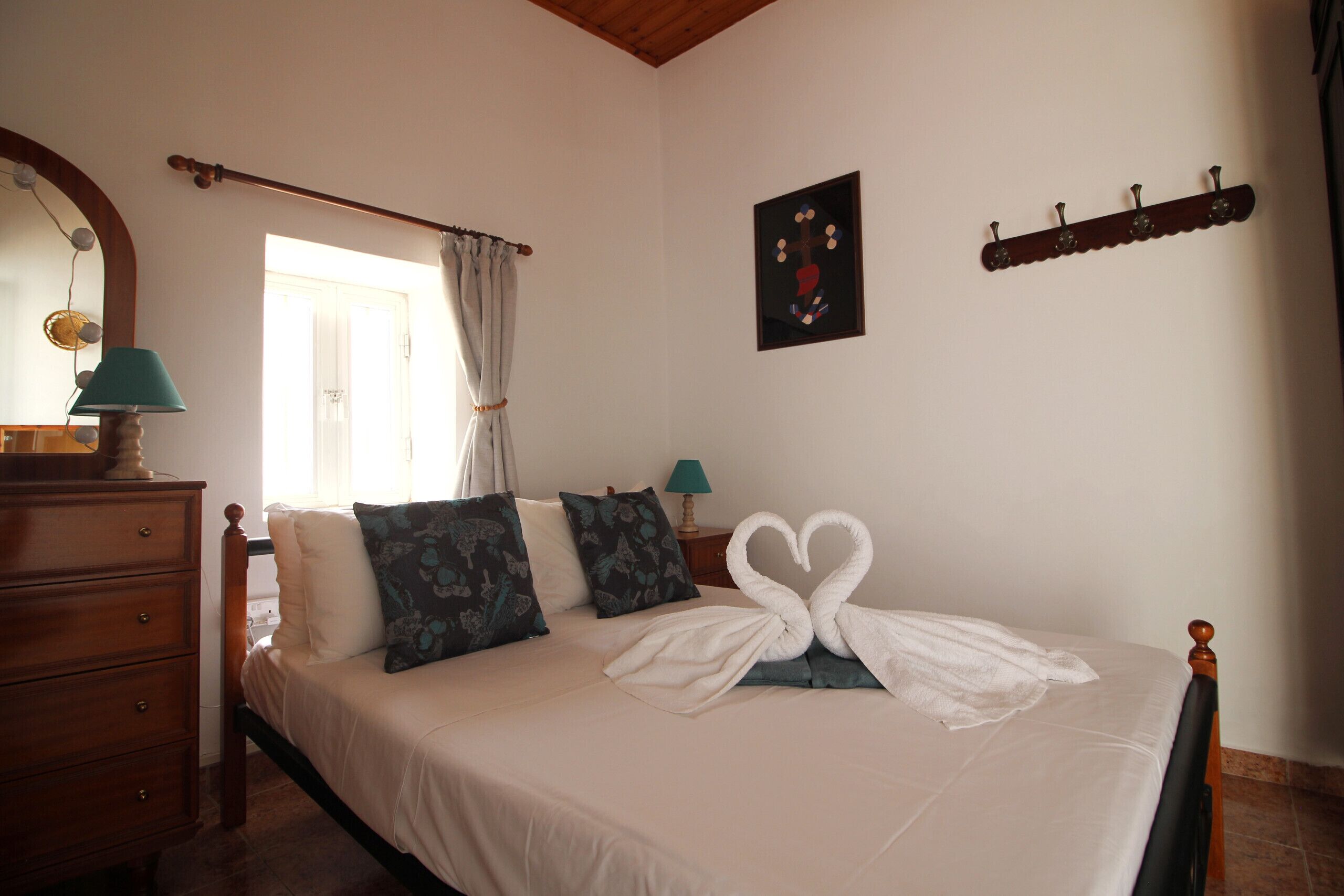 Une chambre d’hôtel comprenant un lit, une commode, un miroir, une fenêtre avec des rideaux, un tableau accroché au mur et un porte-manteau.