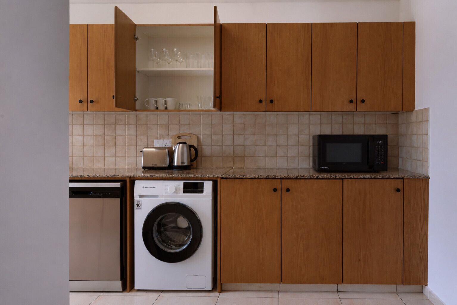 Cozinha compacta com armários de madeira, máquina de lavar, micro-ondas e torradeira.