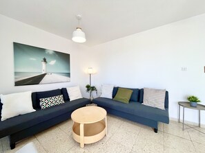 A modern living room with a dark blue sofa, a round wooden coffee table, and a painting of a lighthouse on the wall. - STAY Napa Zotiko Apartment (Ayia Napa)