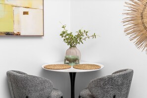 Une pièce avec une table ronde, deux chaises grises, un vase avec des feuilles vertes et une œuvre d’art accrochée au mur.