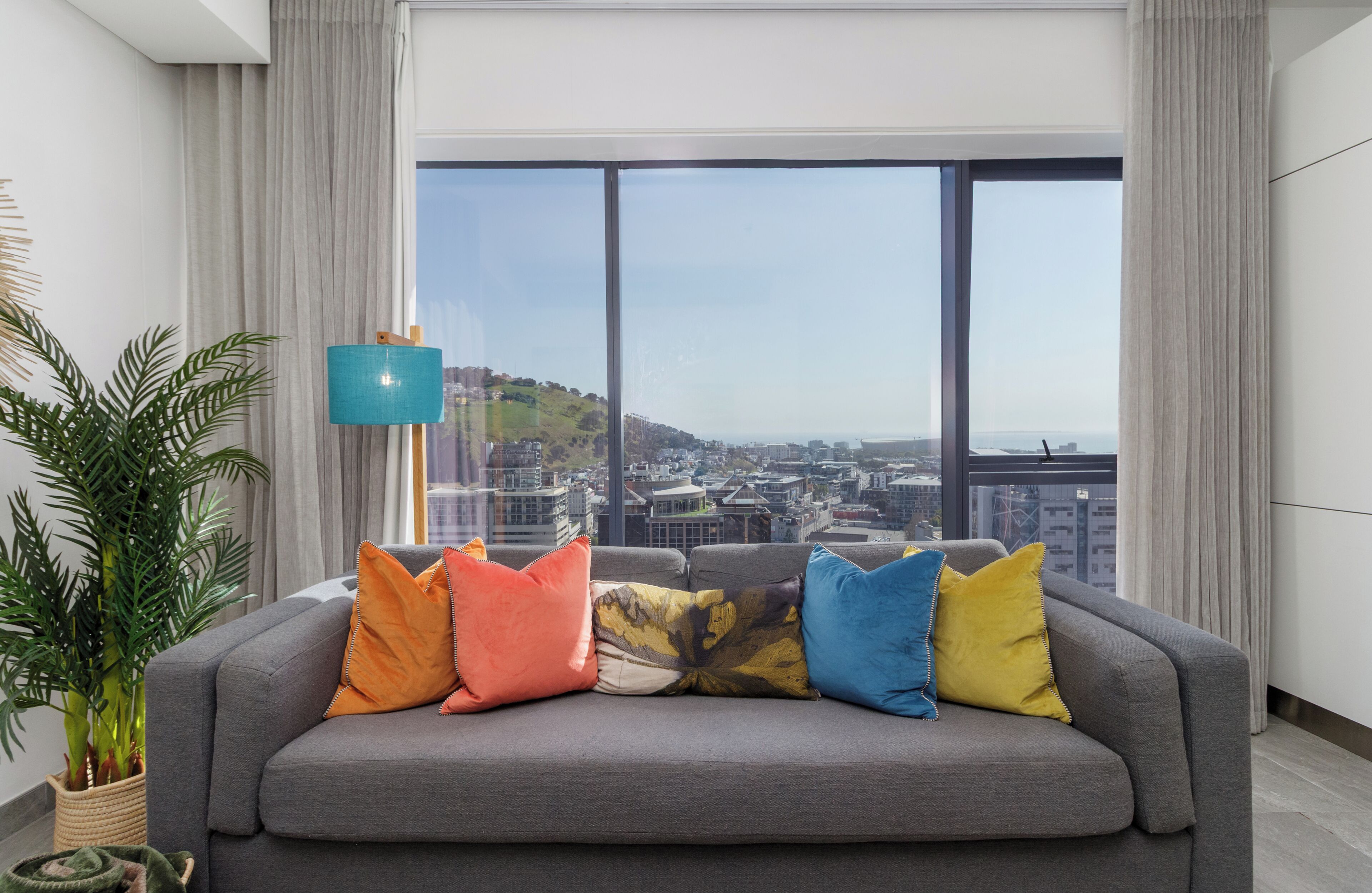 A modern living room with a grey sofa, colorful cushions, a large window with a city view, and a plant.