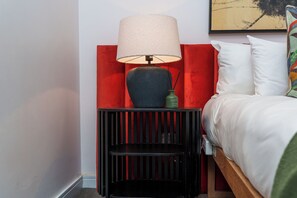 Ein Bett mit weißer Bettwäsche, einem roten Kopfteil, einem schwarzen Nachttisch mit Lampe und einer grünen Vase.
