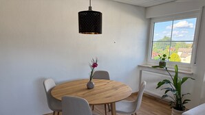 A dining area with a round wooden table, four grey chairs, a vase with a flower, a black pendant light, and a window with a view of greenery and buildings.