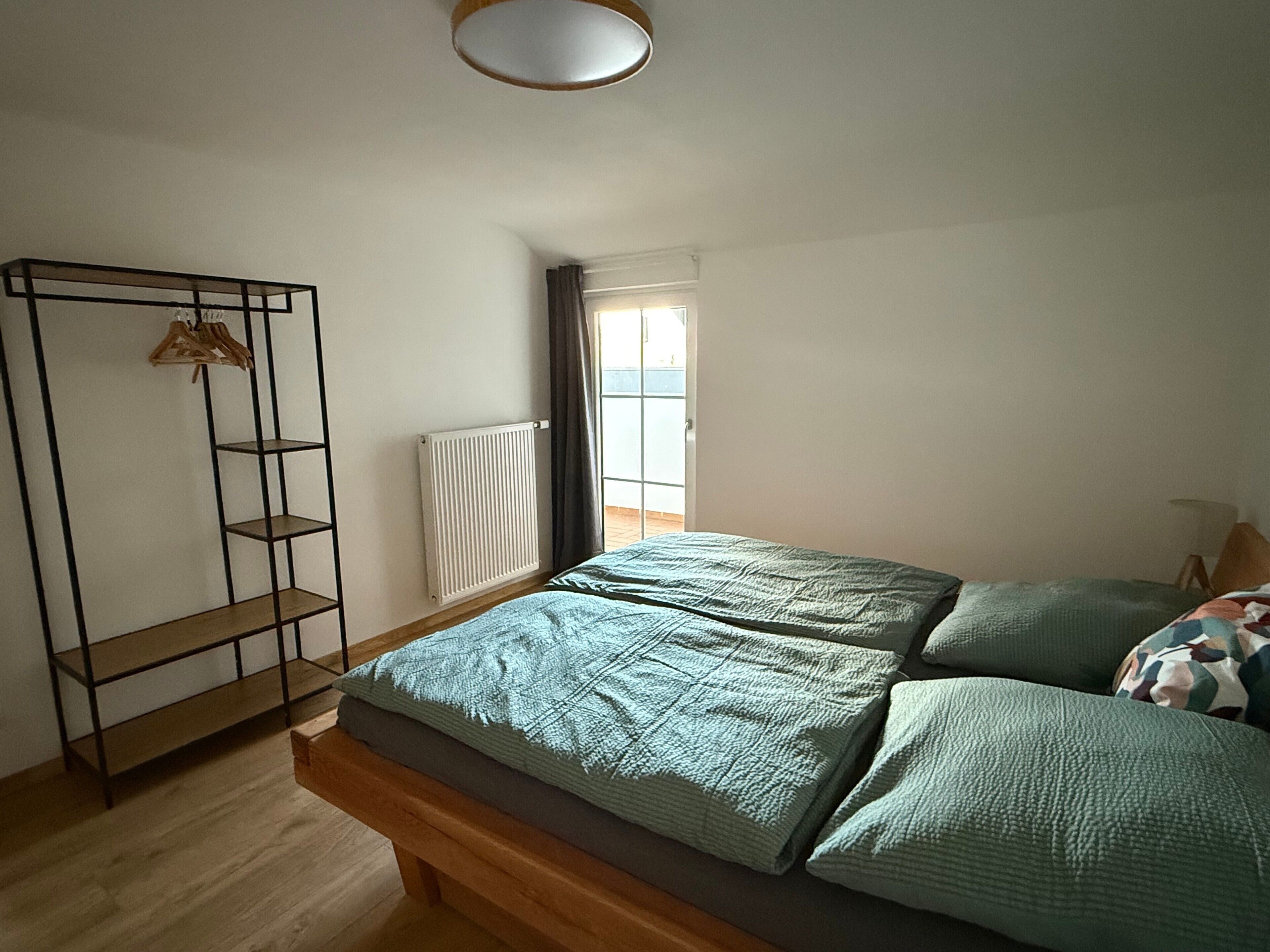 Quarto com cama, um banco de madeira, radiador e janela com cortinas.