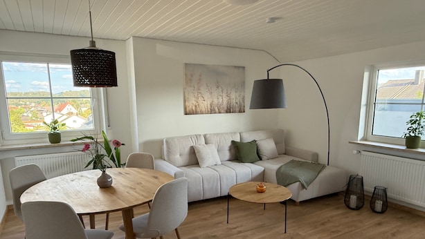 A modern living room with a wooden dining table, a sofa, and a large window with a view of buildings.