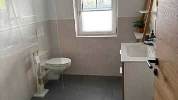 Ein Badezimmer mit Toilette, Waschbecken und Dusche. Über der Toilette befindet sich ein Fenster.