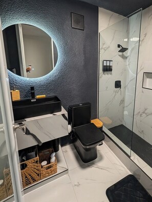 A modern bathroom with a marble floor, a black toilet, a glass shower enclosure, and a large mirror with a circular light. - 2-bedroom with JACUZZI and WATER BACKUP in charming Carolina (Carolina)