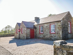 A stone building with a red door and windows, a paved courtyard, and a stone wall. - The Mummers Studio Apartment 1 (Enniskillen)