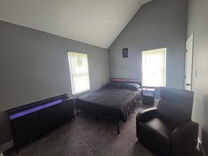 Un dormitorio con una cama, una silla, un aparador y dos ventanas con persianas.