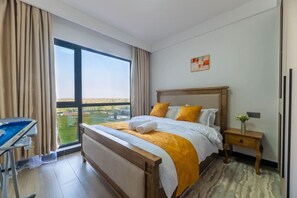 A hotel room with a large bed, a bedside table, a desk, and a view of the outdoors. - The Safari view Gateway | 10 Min JKIA | 5 Min SGR (Nairobi)