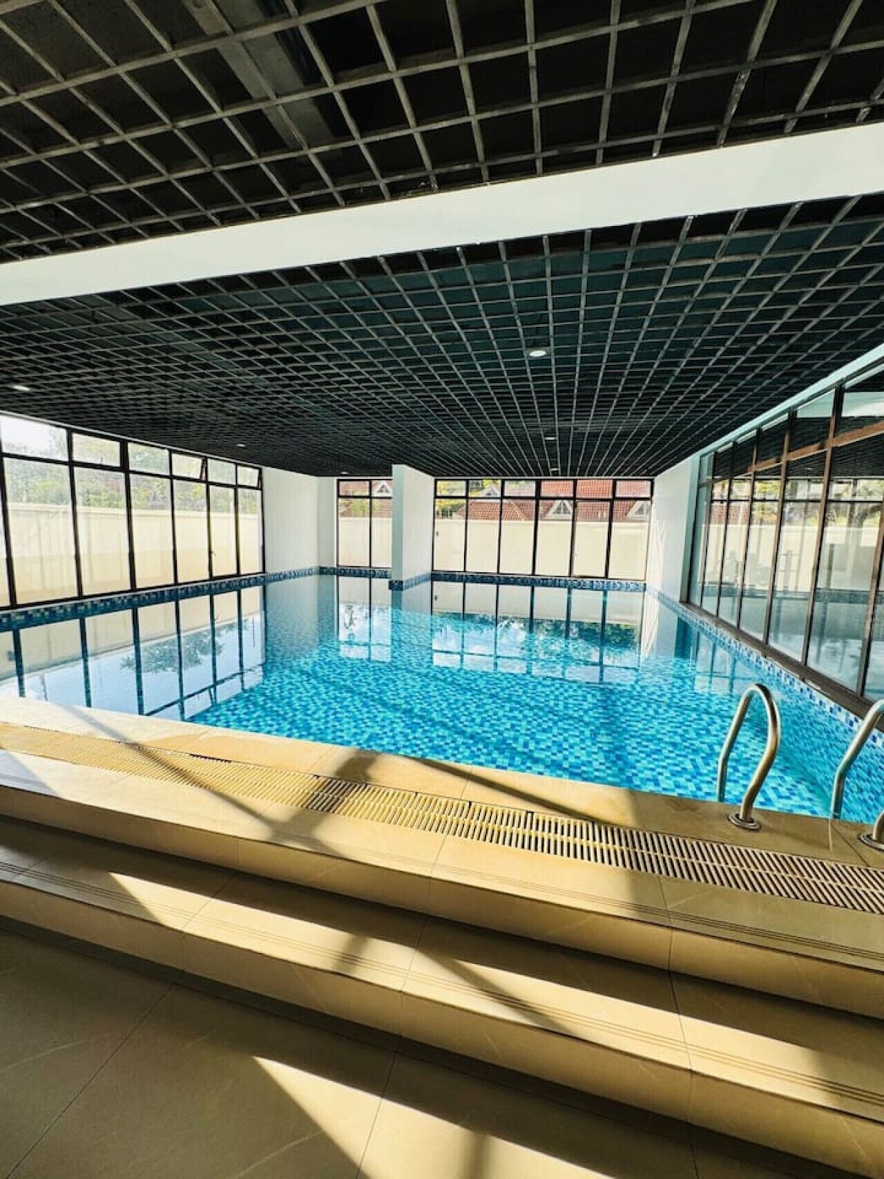 Una piscina interior con paredes de vidrio transparente, piso de baldosas y techo con diseño de cuadrícula.