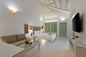 Una habitación de hotel moderna con sofá, una mesa de centro de vidrio con fruta, una cama con contraventanas verdes, un ventilador de techo y un televisor de pantalla plana.