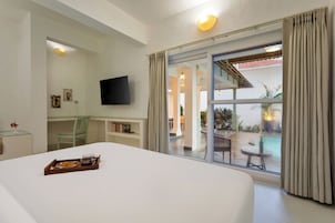 Una habitación de hotel con cama, escritorio, televisor, estantería y vista a un área exterior con piscina y mobiliario de patio.