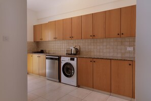 A kitchen with wooden cabinets, a dishwasher, a washing machine, and a countertop with a kettle. - 160 LCA - Stay Sunshine City Apartment (Larnaca)