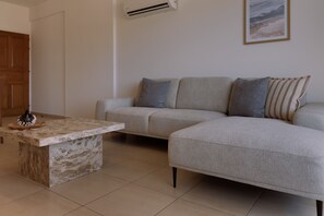 A living room with a sectional sofa, a marble coffee table, and a wall-mounted air conditioner. - 160 LCA - Stay Sunshine City Apartment (Larnaca)