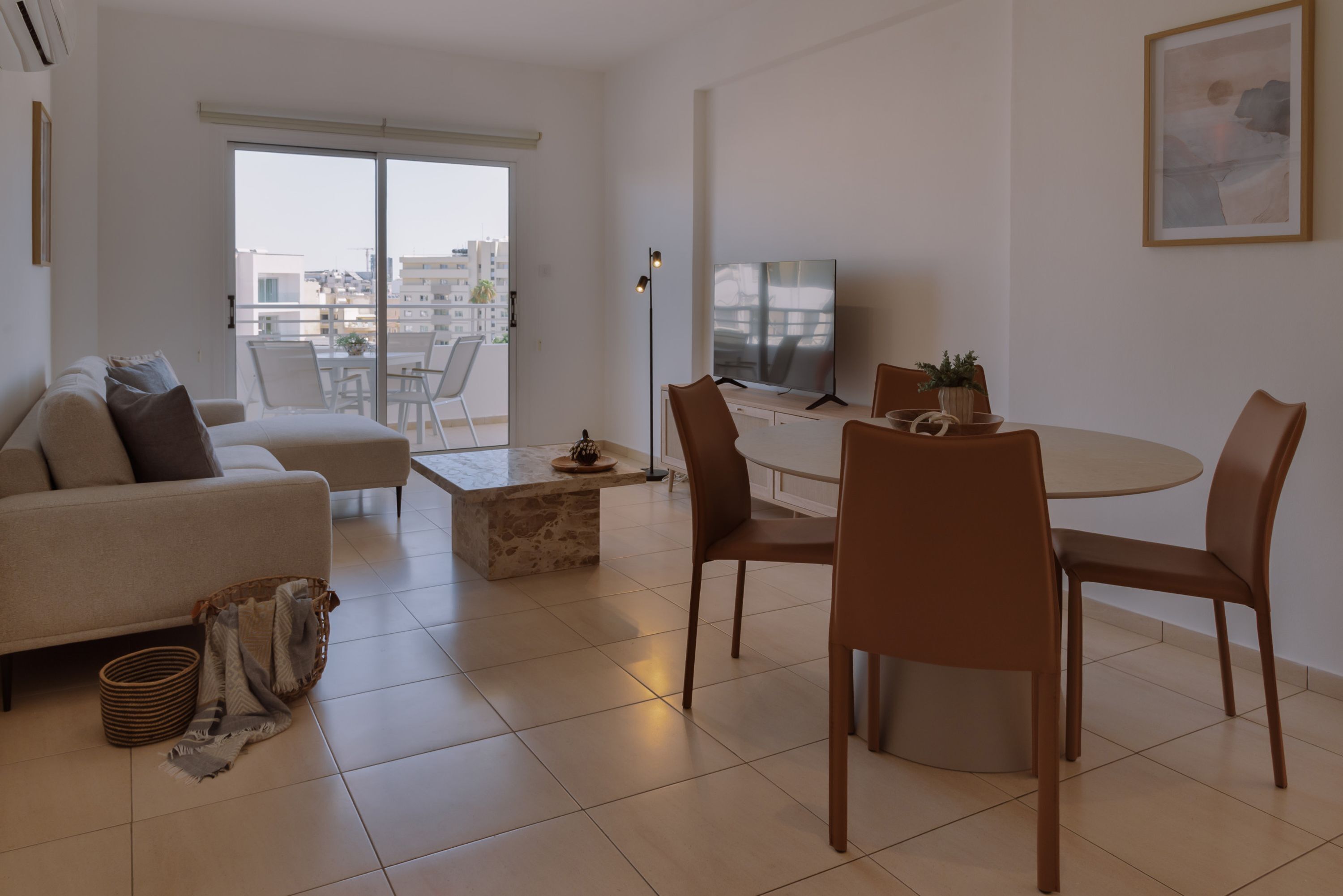 A modern living room with a sofa, a dining table with chairs, a flat-screen TV, and a balcony with a view of buildings.