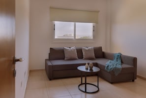 A room with a sofa, a small round table, and a window with a view of buildings. - 160 LCA - Stay Sunshine City Apartment (Larnaca)