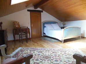A bedroom with a wooden ceiling, a bed with a blue headboard, a wooden dresser, a chair, a mirror, and a patterned rug. - Charming country house on the outskirts of the Périgord (GAVAUDUN)