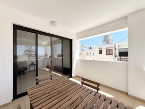 A balcony with a wooden bench, a sliding glass door leading to a room with a chair and a small table, and a view of neighboring buildings. - 161 AN - Stay Napa Zotiko Apartment (Ayia Napa)