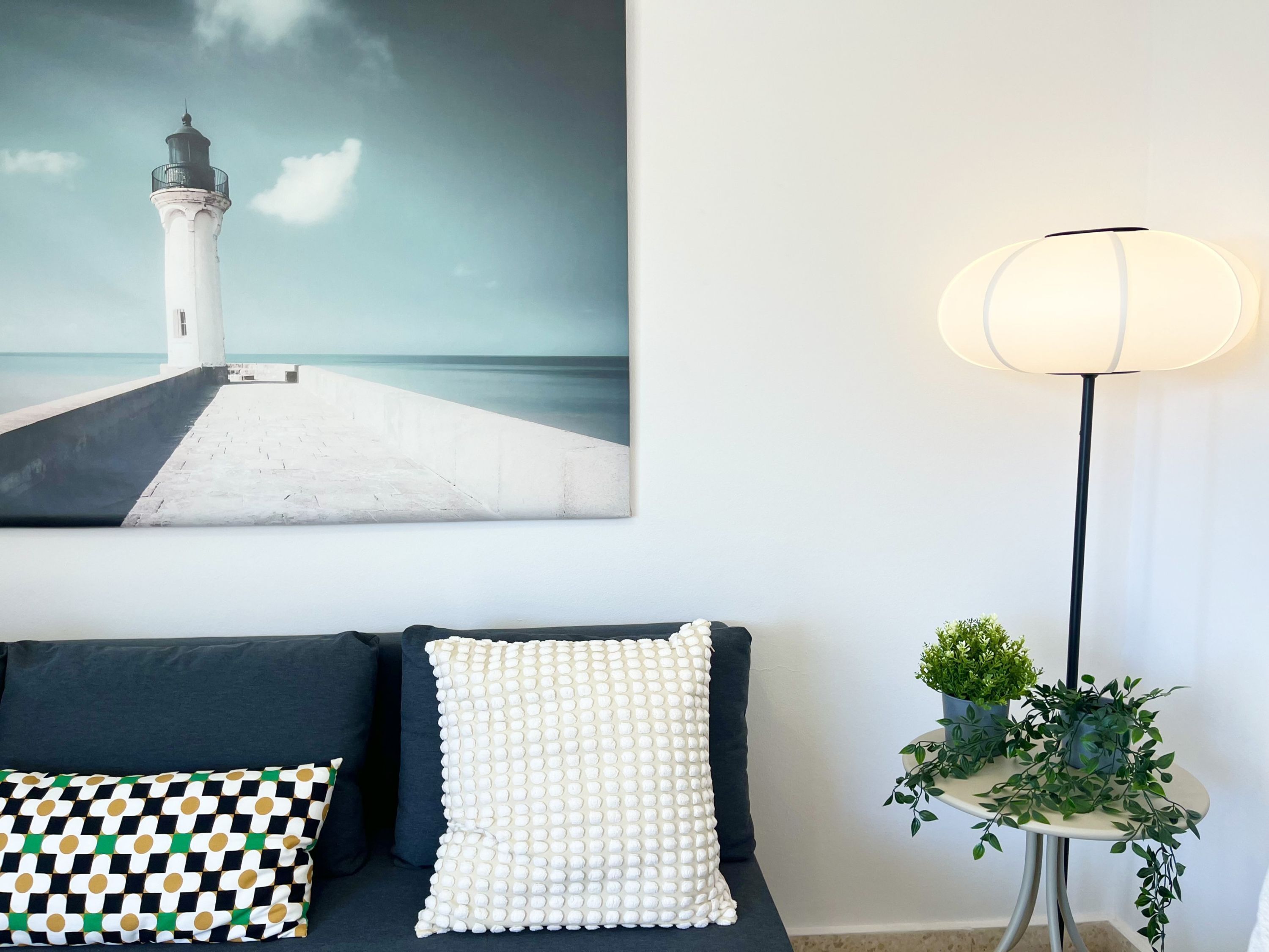 A room with a dark-colored sofa, a white pillow with a textured pattern, a patterned pillow, a wall-mounted painting of a lighthouse, and a floor lamp with a white shade.