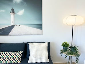Ein Zimmer mit einem dunkelbraunen Sofa, einem weißen Kissen mit strukturierter Oberfläche, einem gemusterten Kissen, einem an der Wand hängenden Gemälde eines Leuchtturms und einer Stehlampe mit weißem Lampenschirm.