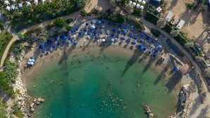 An aerial view of a beach with numerous blue umbrellas and lounge chairs arranged in a semi-circular pattern around a central water body. - 155 PRO - Stay Prime View Apartment (Paralimni)
