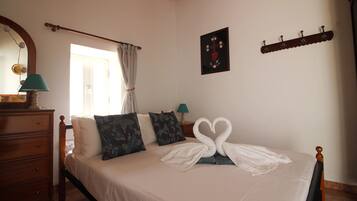 Un dormitorio con cama, cómoda, espejo, ventana con cortinas, un cuadro en la pared y perchero.