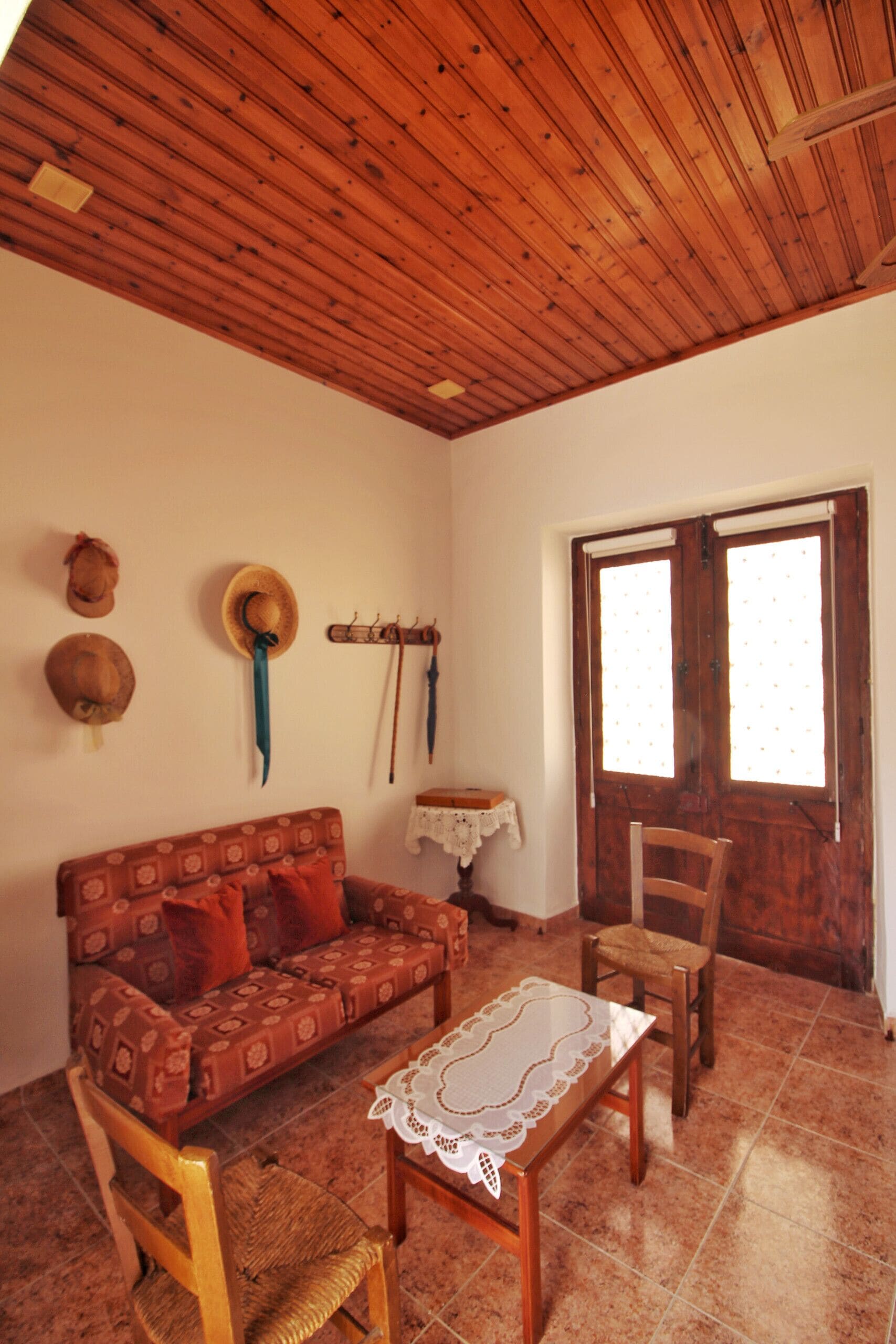 Una sala de estar con techo de madera, un sofá, un sillón, una mesita y un ventanal con cortinas.