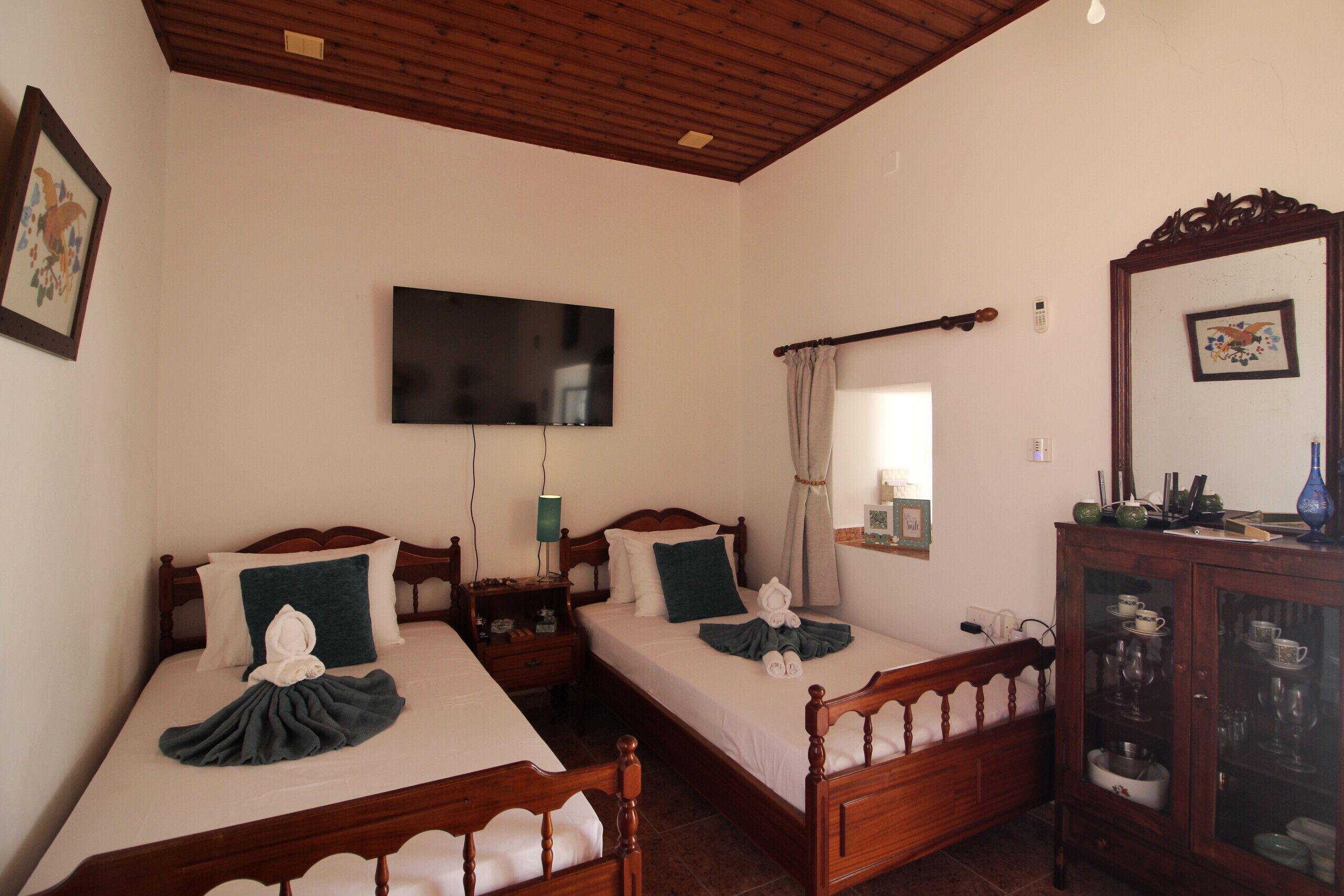 Habitación de hotel con dos camas de madera, un televisor de pantalla plana, una mesita de noche con lámpara, un espejo y un mueble con cristalería.