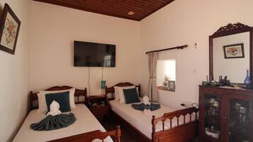 Habitación de hotel con dos camas de madera, un televisor de pantalla plana, una mesita de noche con lámpara, un espejo y un mueble con cristalería.