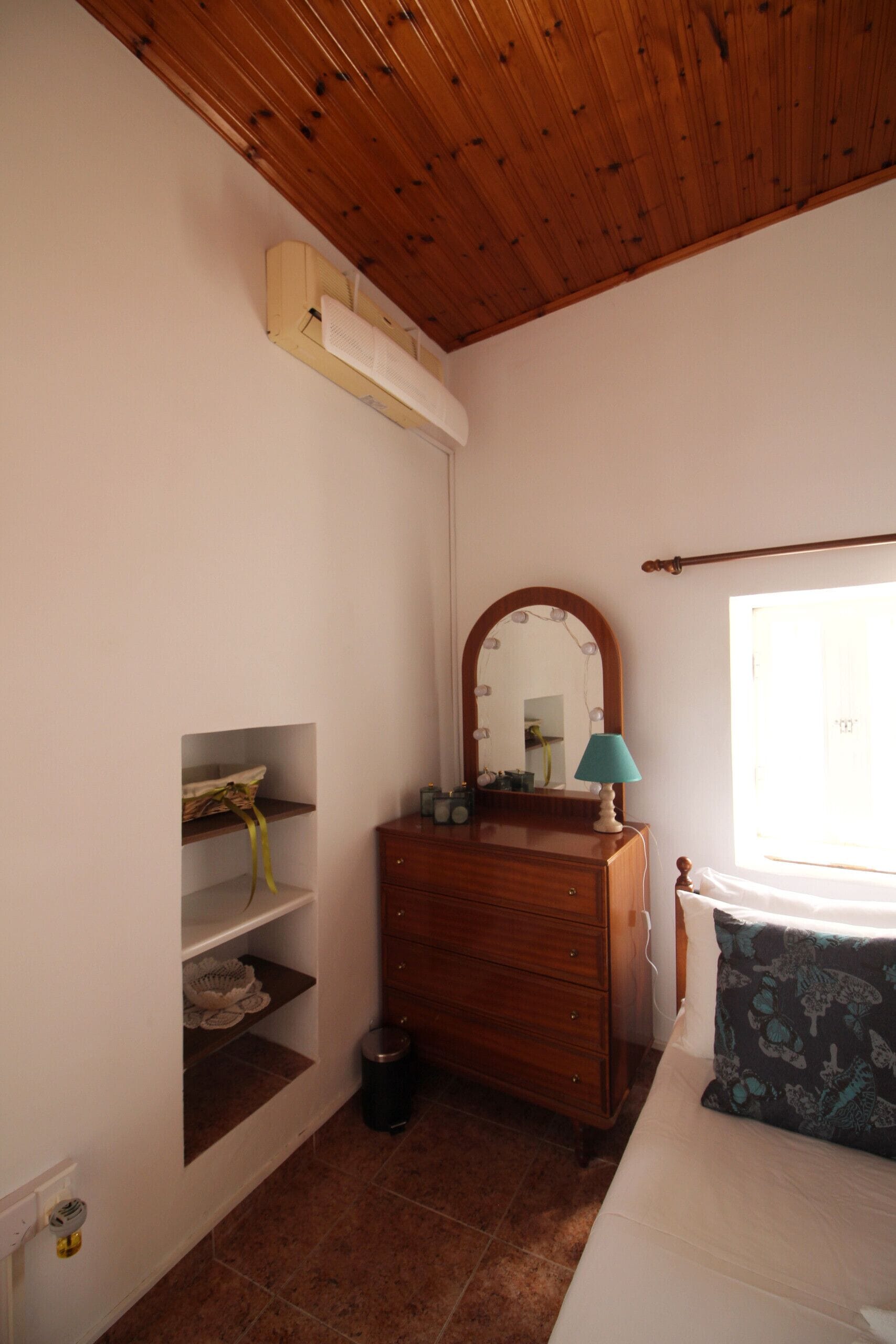 A bedroom with a wooden ceiling, a dresser with a mirror, a lamp, and a window.