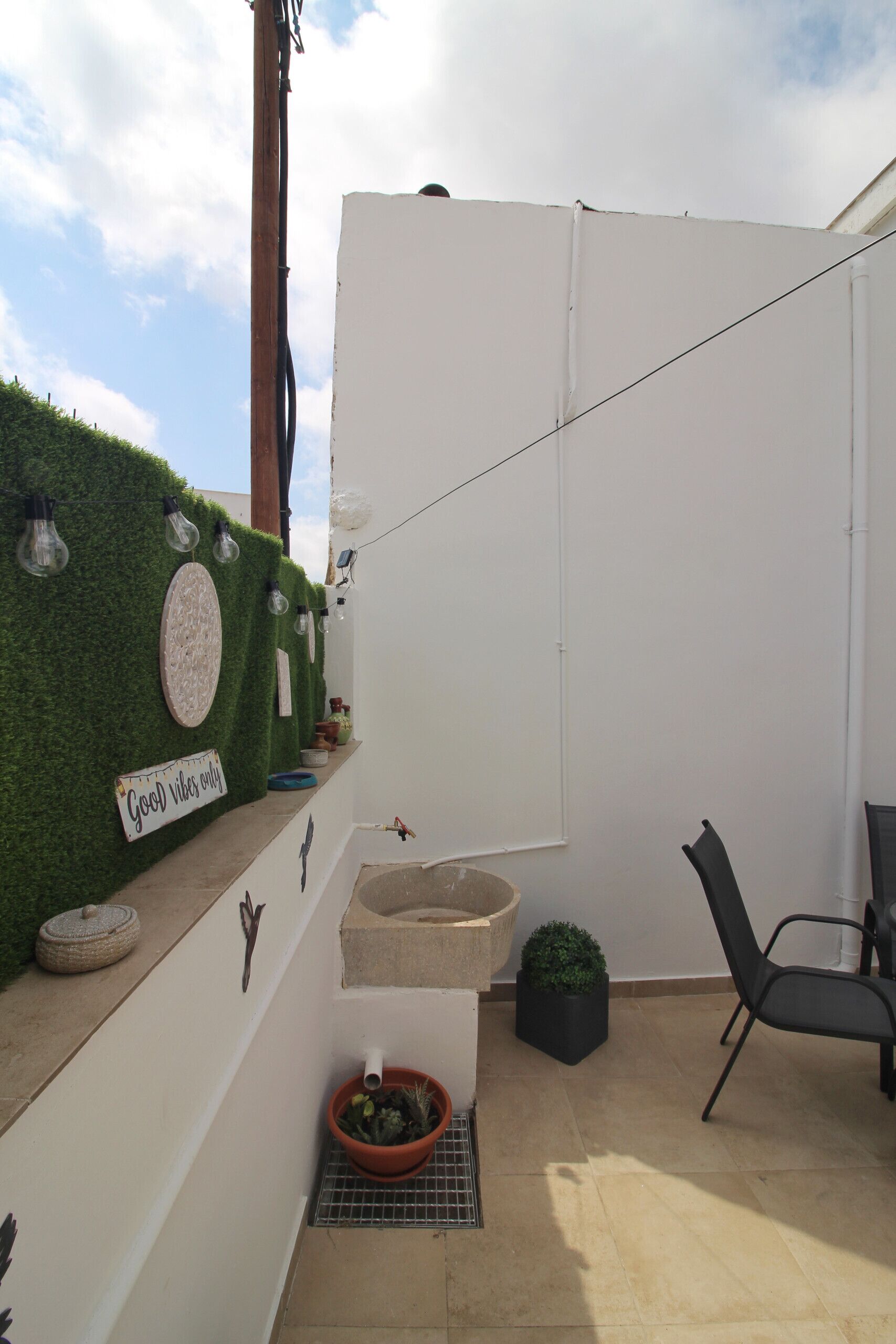 Una terraza en la azotea con un muro blanco, un lavamanos pequeño, una silla y una planta en maceta.