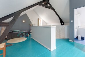 Una habitación con techo inclinado, una cama con colcha estampada, una mesita pequeña y un panel blanco.