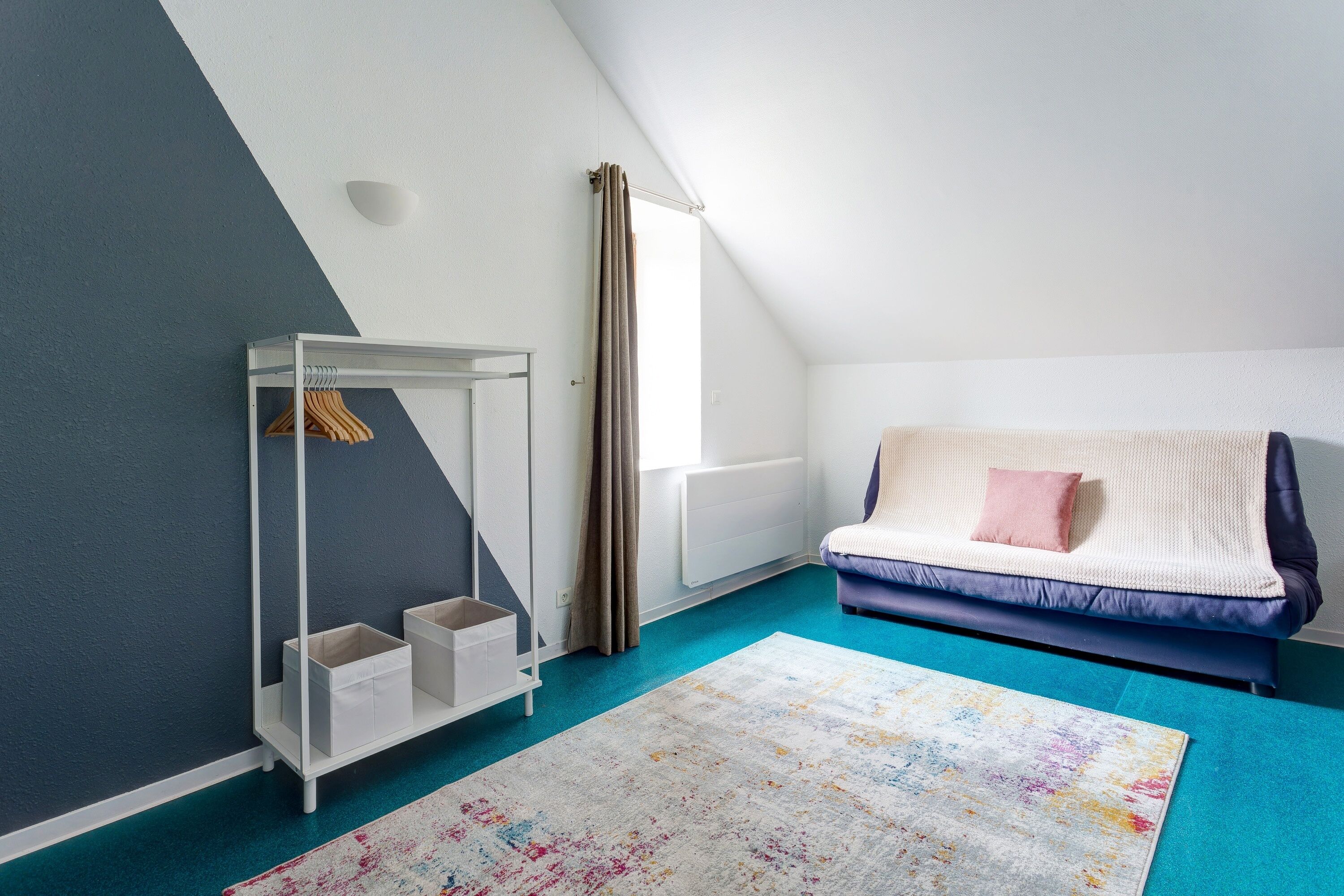 Ett rum med ett sluttande tak, ett vitt element, en blå soffa med en rosa kudde, ett vitt förvaringsskåp med upphängda kläder och en färgglad matta.