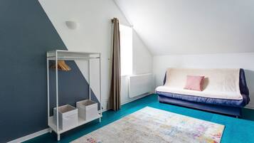 Ett rum med ett sluttande tak, ett vitt element, en blå soffa med en rosa kudde, ett vitt förvaringsskåp med upphängda kläder och en färgglad matta.