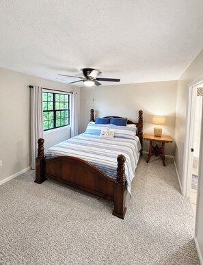 A bedroom with a wooden bed, a ceiling fan, a window with curtains, a bedside table with a lamp, and a carpeted floor. - The Lookout at Lakeshore Estates (Hardin County)