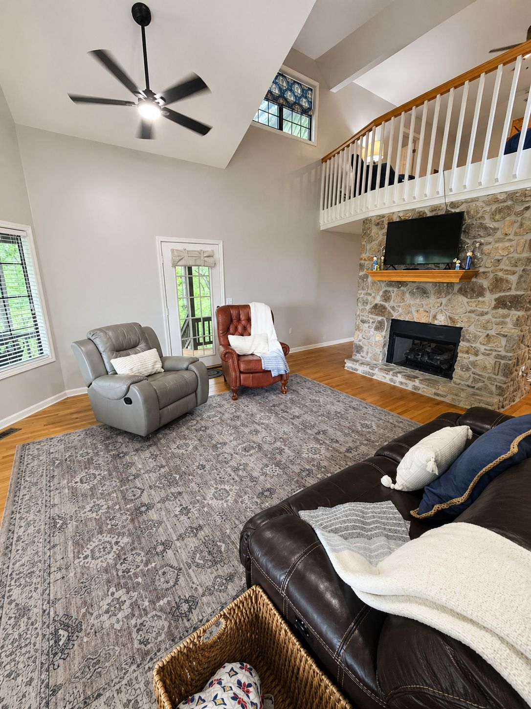 A spacious living room with a fireplace, a stone wall, a ceiling fan, and a large window.